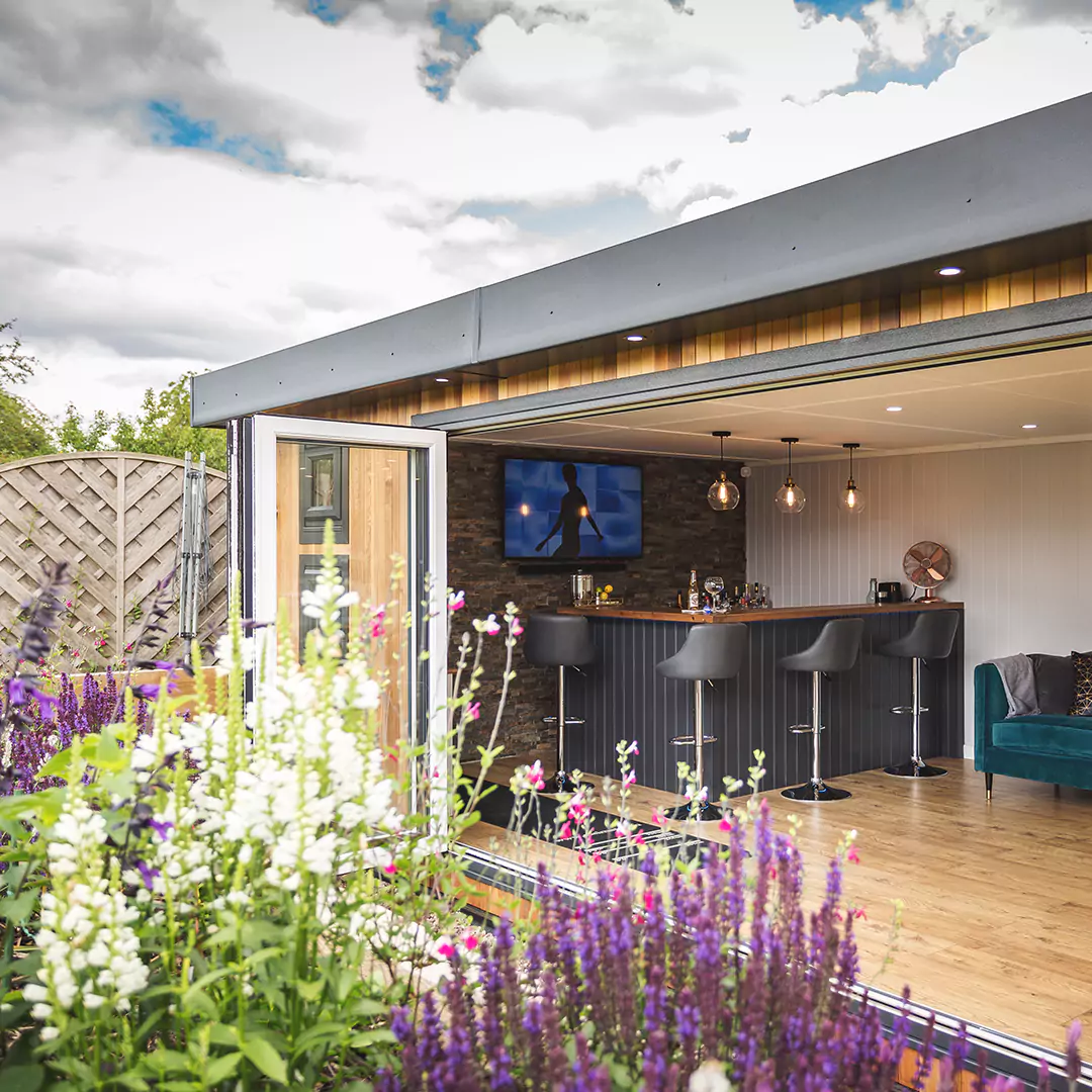 a garden area with flowers and a large timber garden bar building in the background with open bifolds