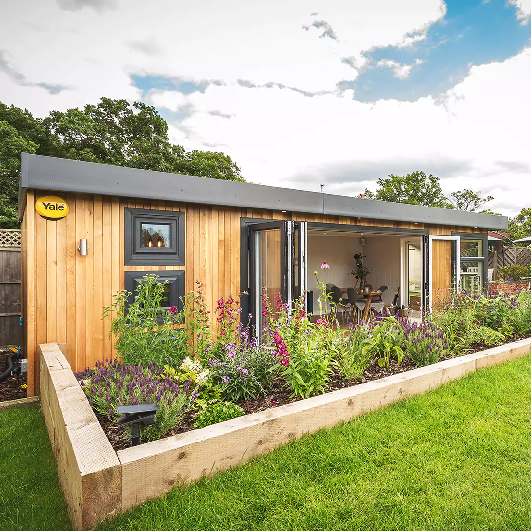 external shot of a cedar clad garden bar with bi-fold doors and raised planter with english flowers