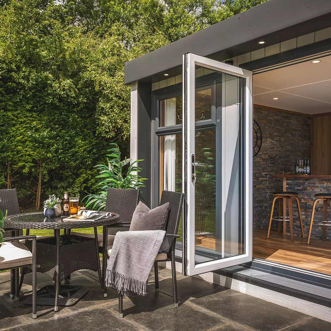 Marley Cabin Master Building with open Bi-Fold Doors on patio area with rattan table and chairs and foliage in background