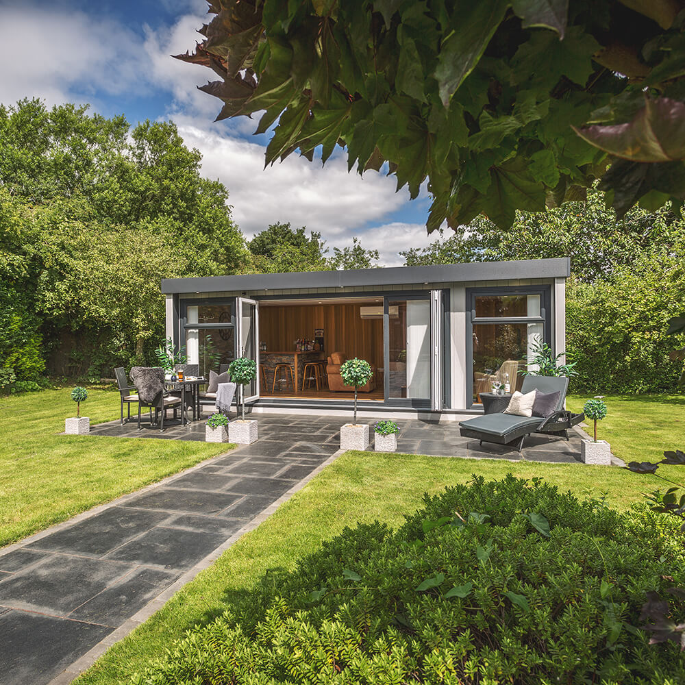 Marley Cabin Master Building with open Bi-Fold Doors on patio area with rattan table and chairs and foliage in background with large garden path and green lawn area