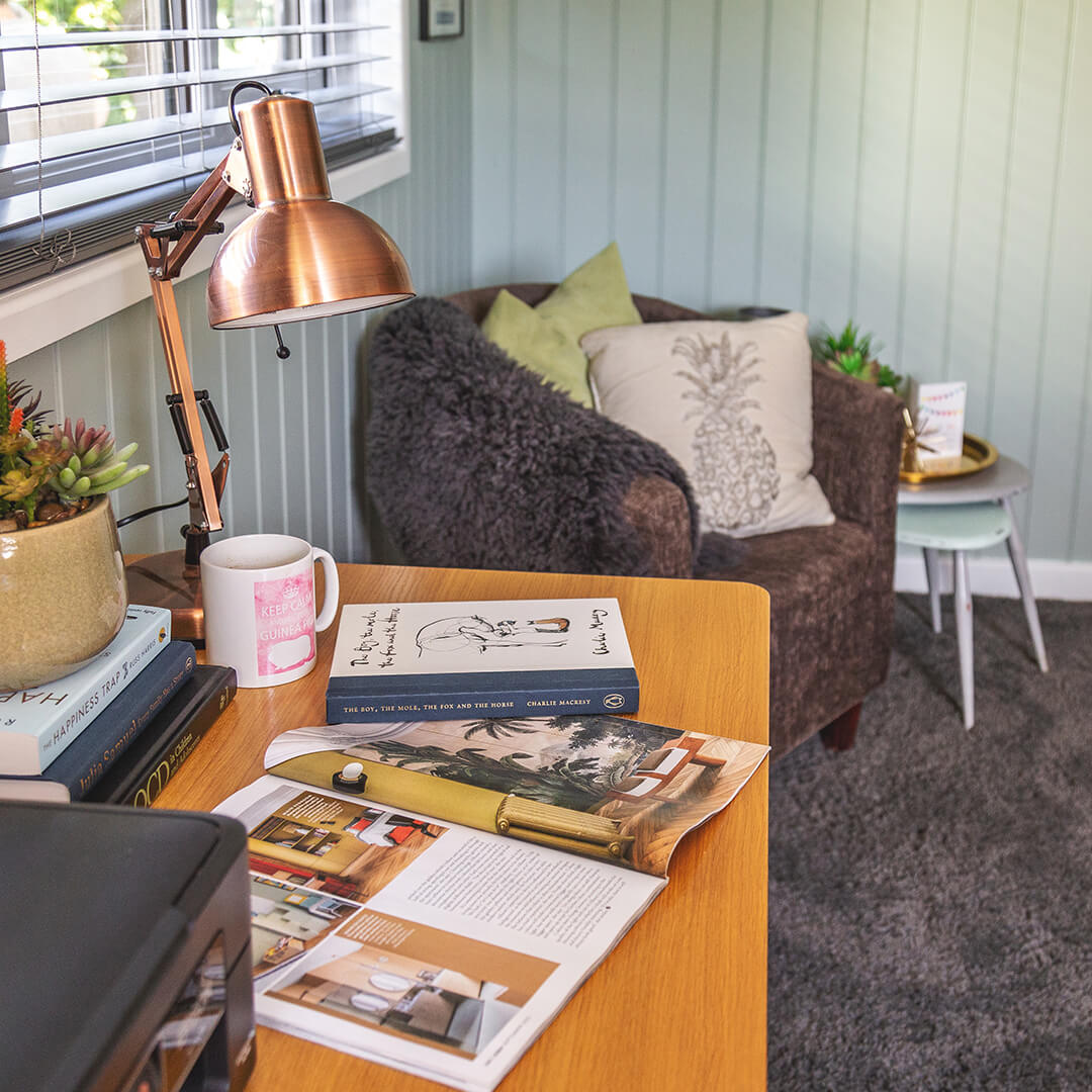 Internal shot of Cabin Master Small Garden Office Pod with desk, lamp, book and magazines in foreground and furry brown armchair and carpet in background