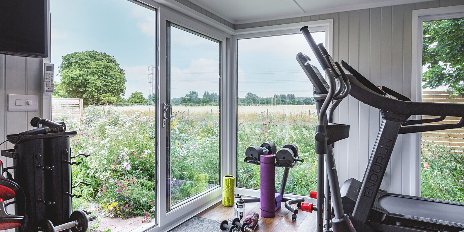 Interior of a garden room gym looking out into a lovely picturesque garden, with gym equipment seen 
