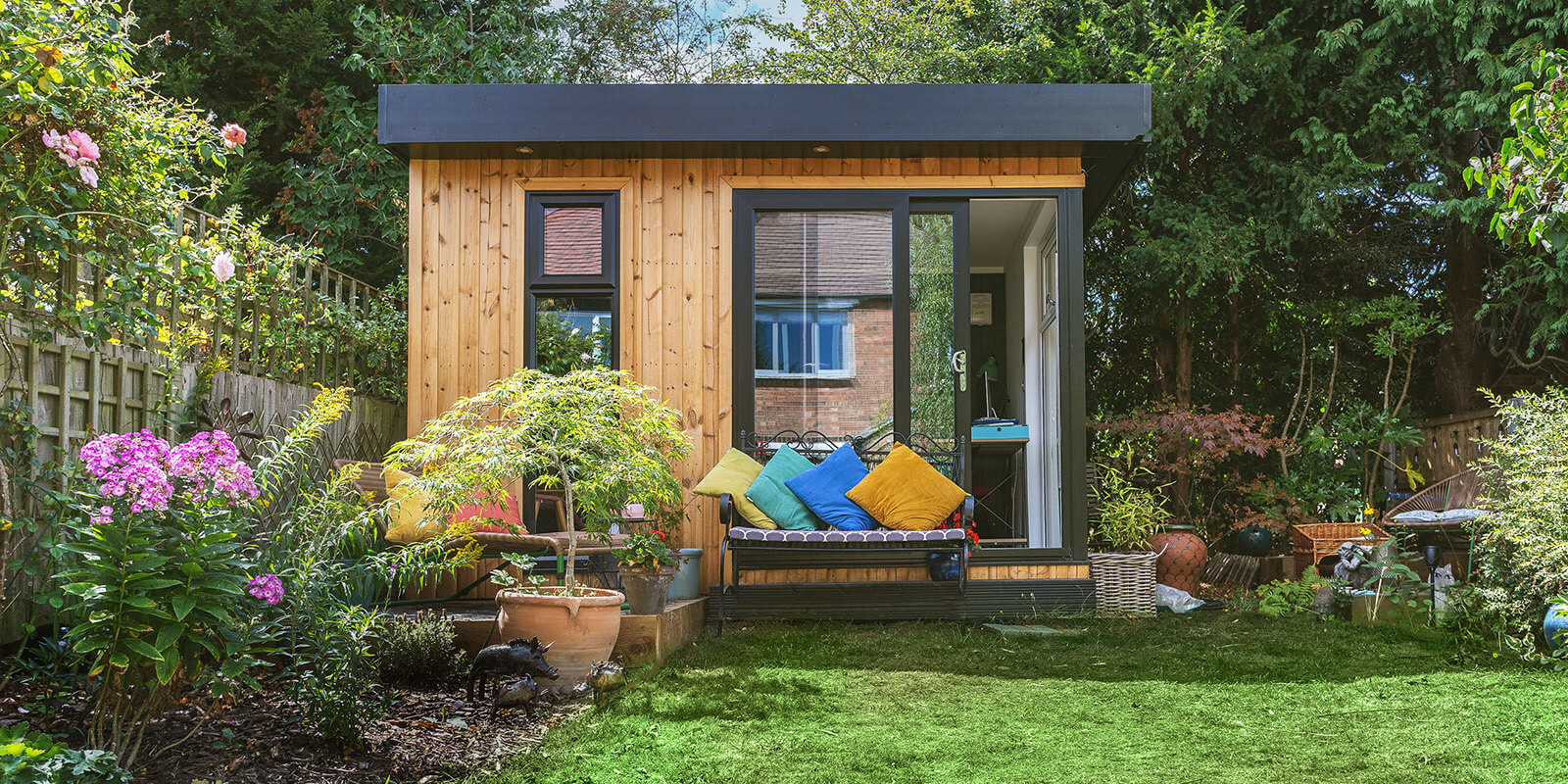 Small insulated garden room in redwood with bright cushions outside 