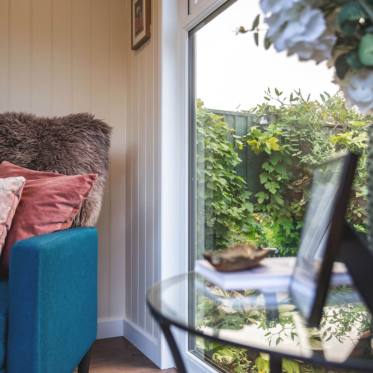 small garden summerhouse with velvet armchair and large glass windows with coffee table and framed photograph on it 