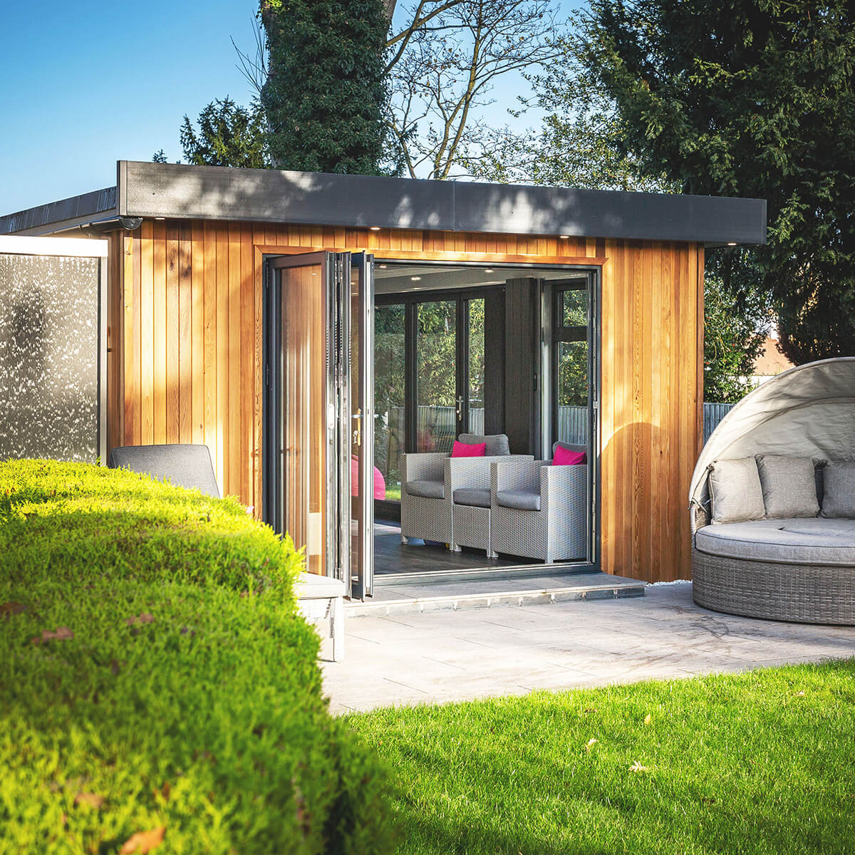 Cedar cabin master garden building with large open bi-fold doors and green lawn area with rattan outdoor furniture