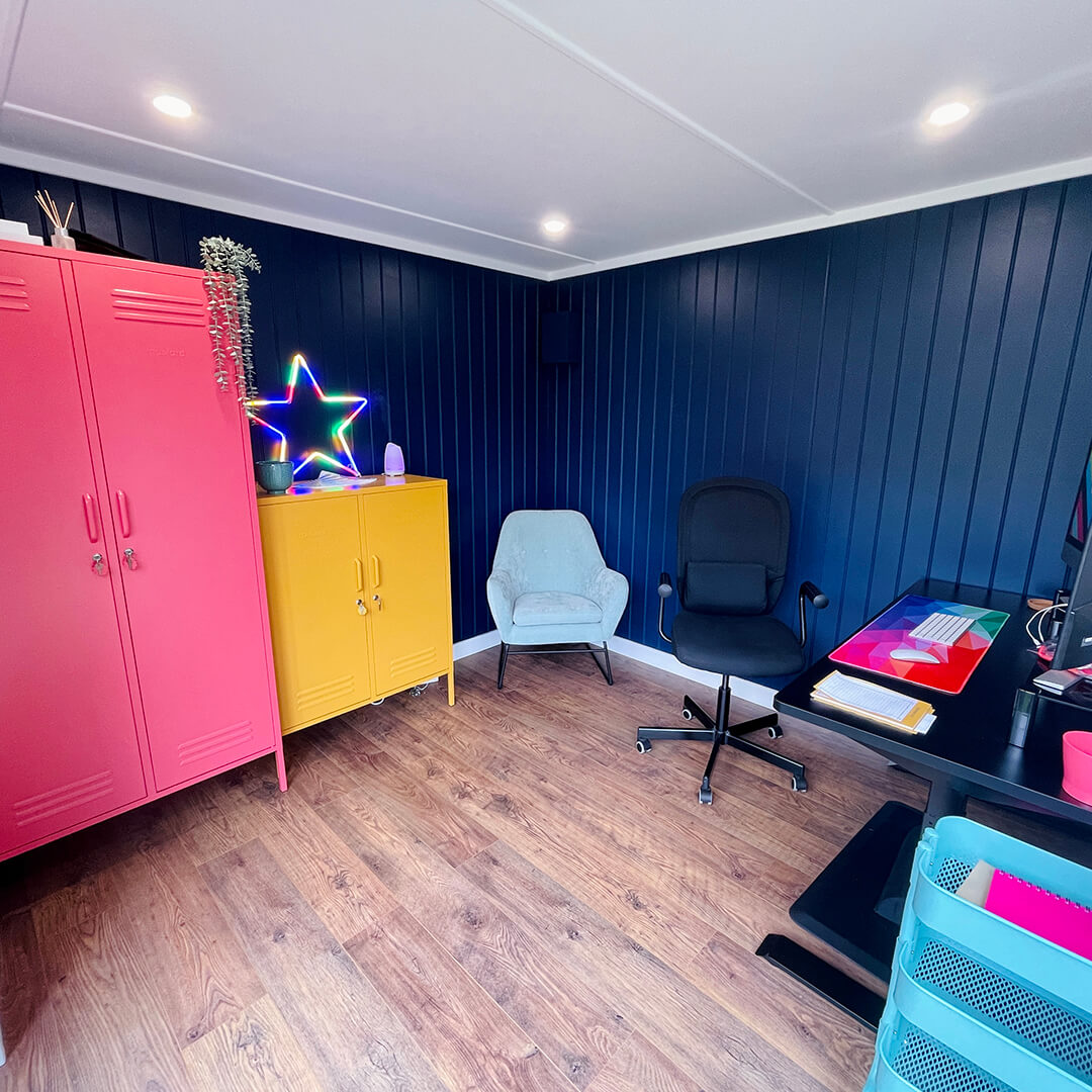 Internal view of Cabin Master Micro Office with navy feature wall and bright yellow and pink cupboards and black desk chair and desk