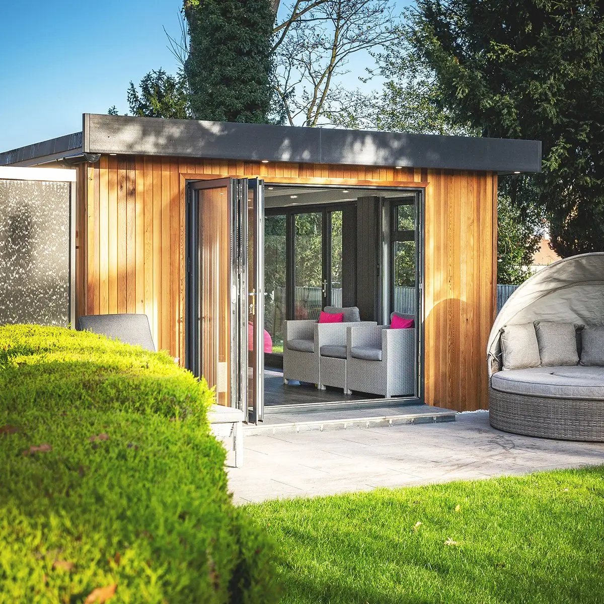 Cedar Garden Room in grassy area with patio and bifold doors and garden furniture and green hedges