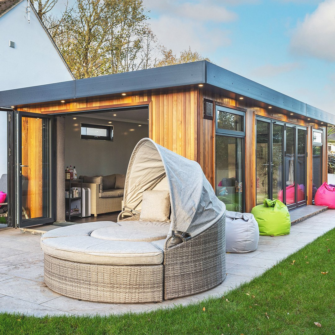 Large swim spa garden building with rattan sofa bed in front of it on large patio area. There are also 3 brightly coloured bean bags leaning against the building. The building has large bifold doors which are open.