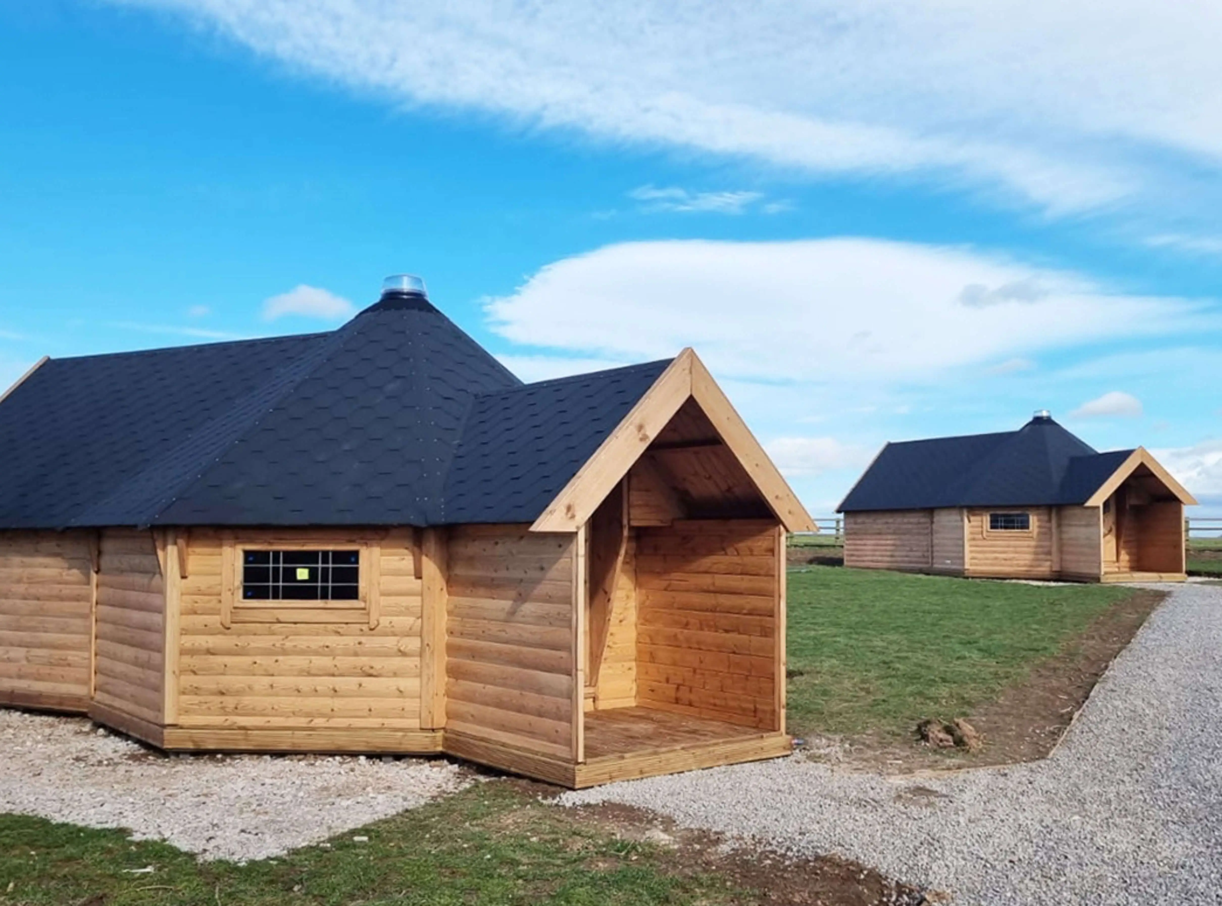 Large Camping Cabins redwood building with black roof tiles and gravel path and grassy area with blue skies