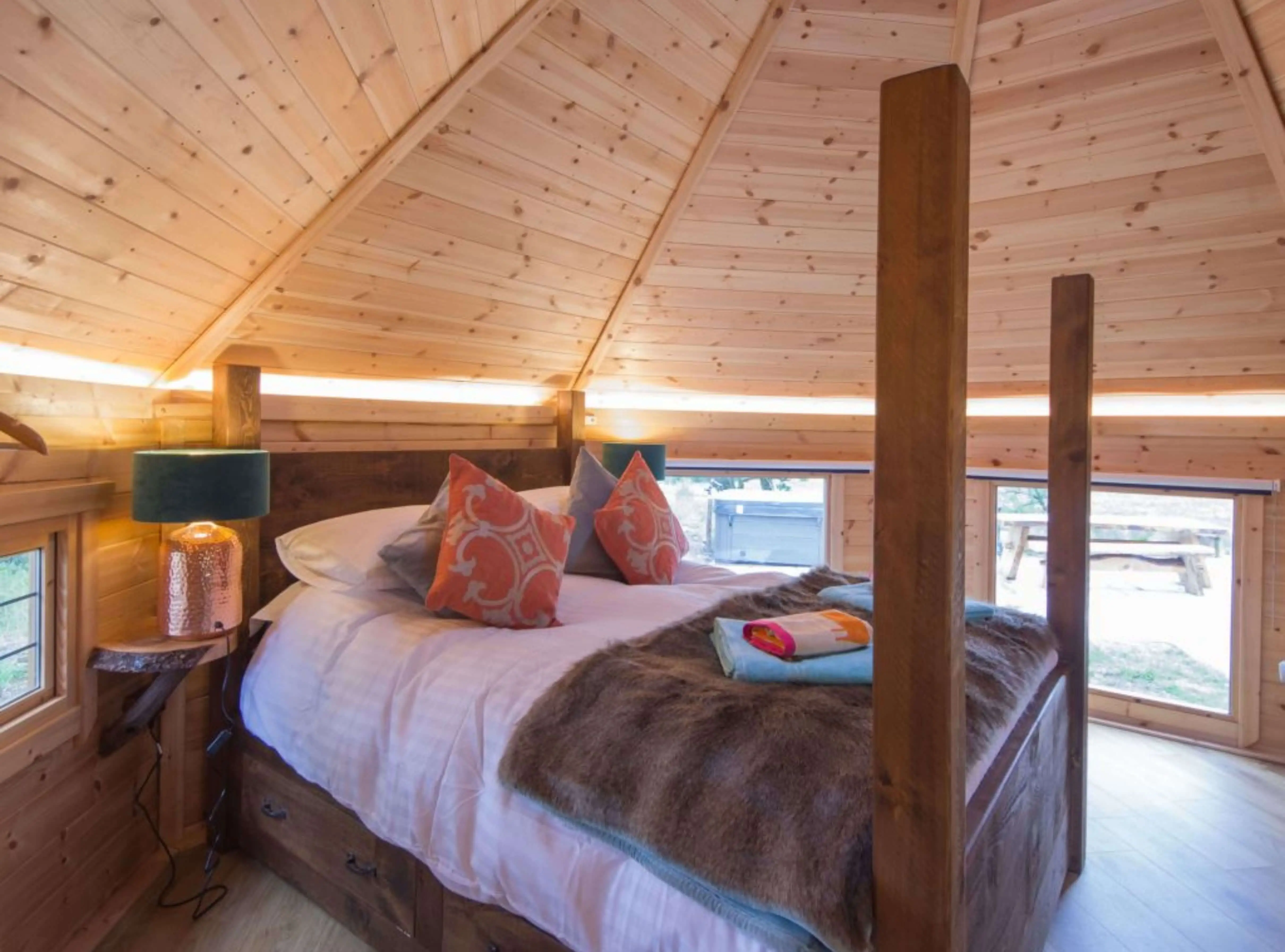 Double bed with faux fur throw and patterned cushions inside large camping cabins redwood building with 4 post bed and led lights