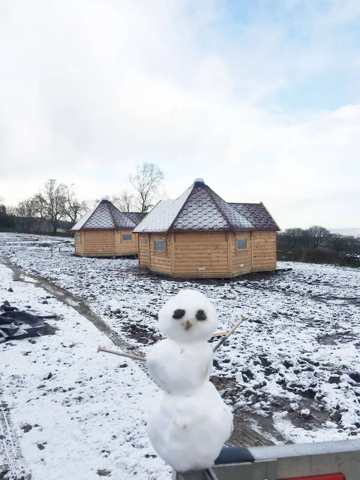 2 red roofed camping cabins with snowman in foreground and snowy pathway and blue sky 