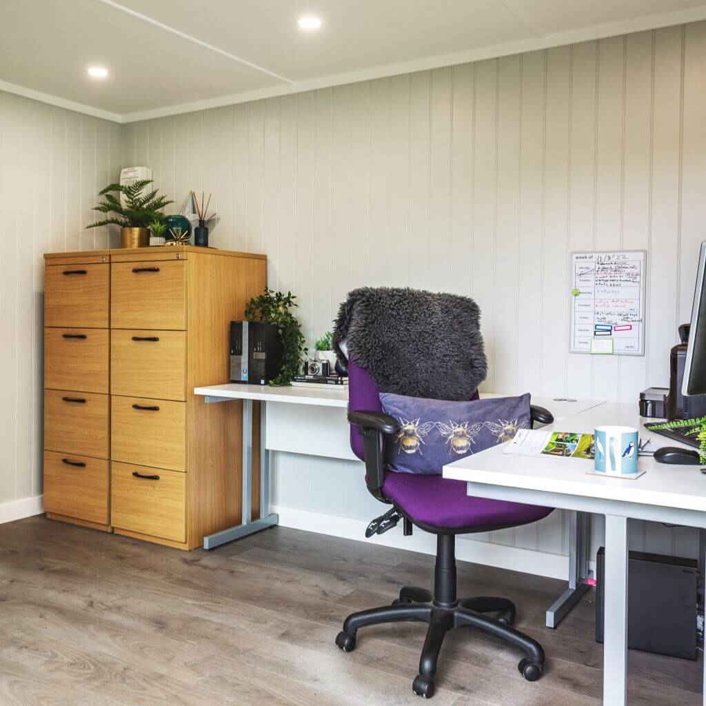 inside view with desk chair and chest of drawers and large white desk with indoor plants and purple sofa cushion with bees on it