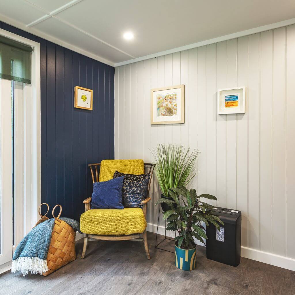 Inside view of corner redwood garden office with large indoor plants and yellow armchair with wall hangings 