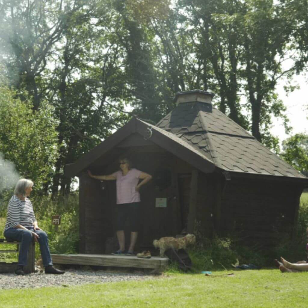 2 women sitting outside eco pod camping cabin building in field with dog and timber steps