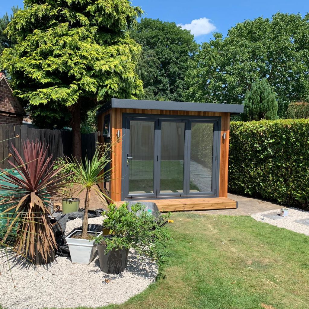 small micro office with bifold doors in lawn area with gravel planter and large oak trees in the background