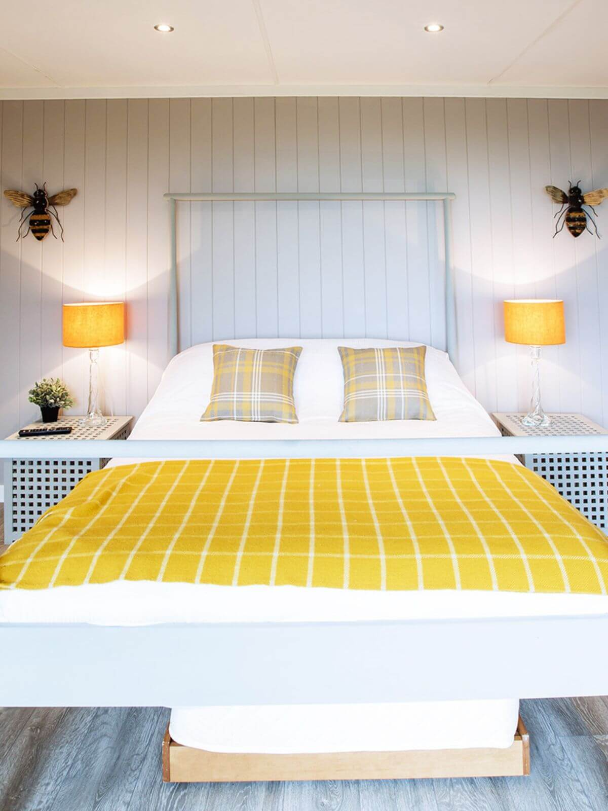inside view of glamping lodge with double bed and spotlights and yellow checked blanket and checked cushions and bumble bee wall hangings and grey flooring and yellow lamp shades
