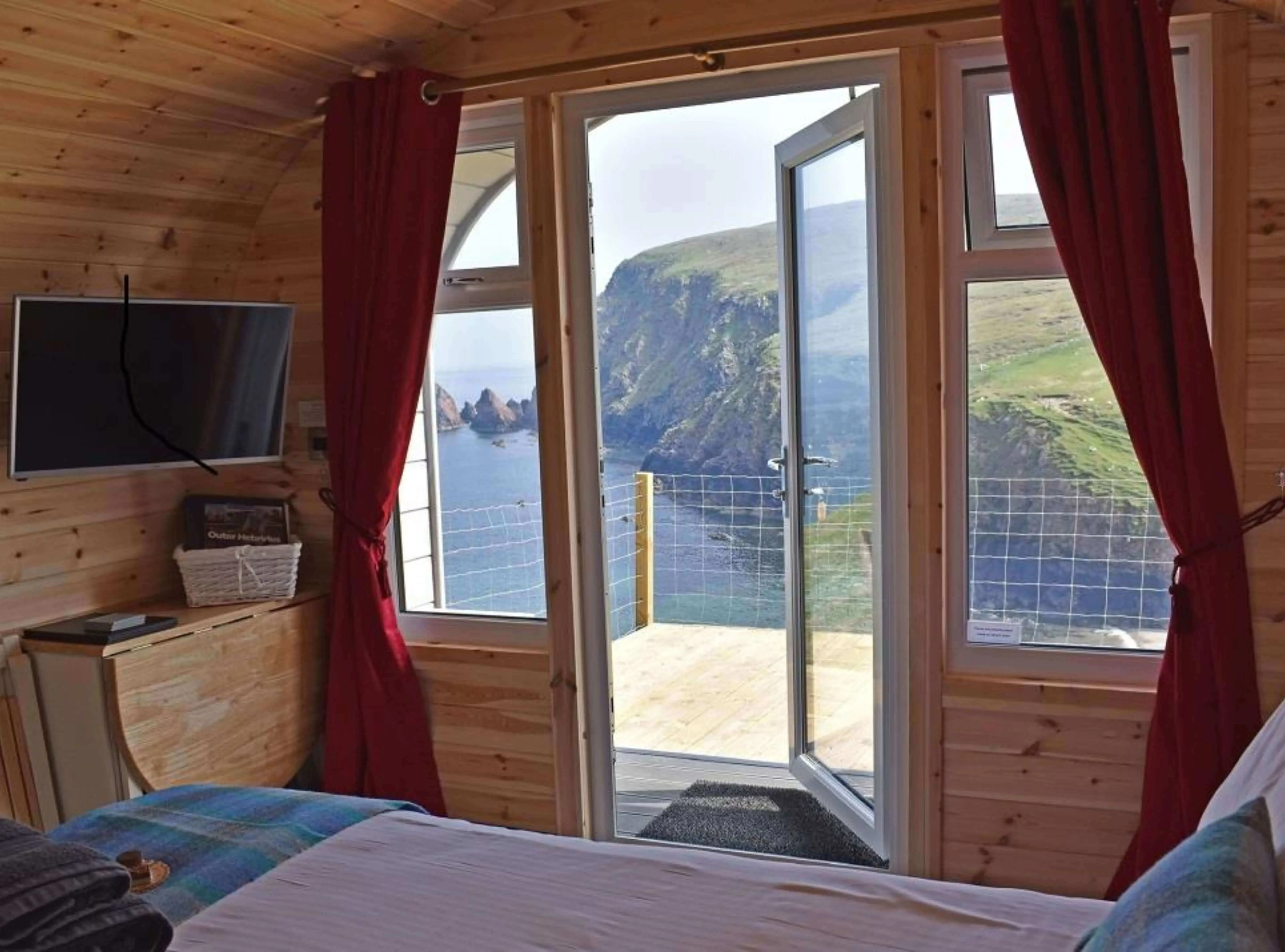inside of camping cabins glamping lodge with flatscreen tv and red curtains and balcony with view of cliffside and sea
