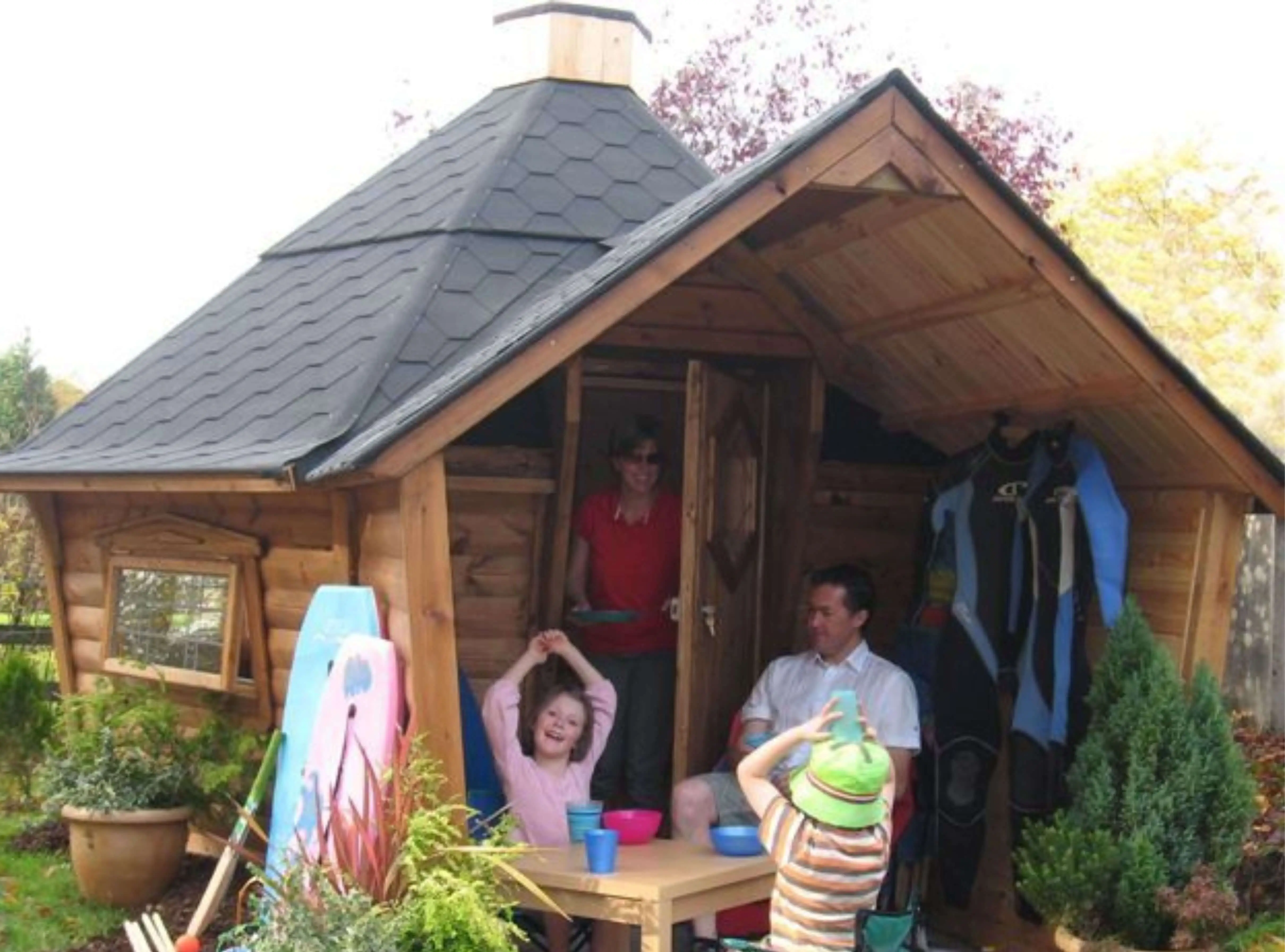 camping cabin with black roof tiles with family seated outside and plants and coats hanging on coat rack